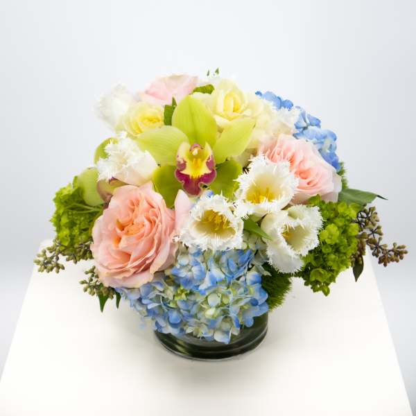 Pastel arrangement of pink roses, white fringed tulips, green orchids, and blue hydrangeas in a glass vase