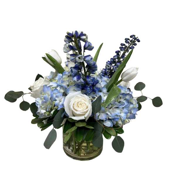 Blue hydrangeas, white roses, white tulips, and blue delphinium arranged in a clear glass cylinder vase
