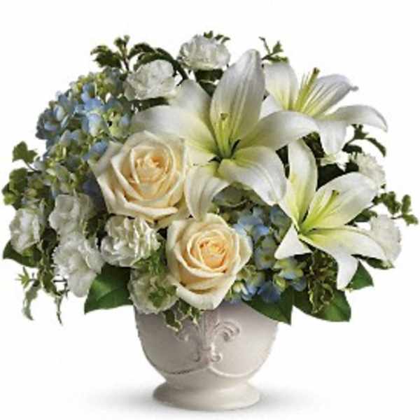 Ivory lilies, cream roses, and white carnations arranged with blue hydrangea in a white ceramic urn vase