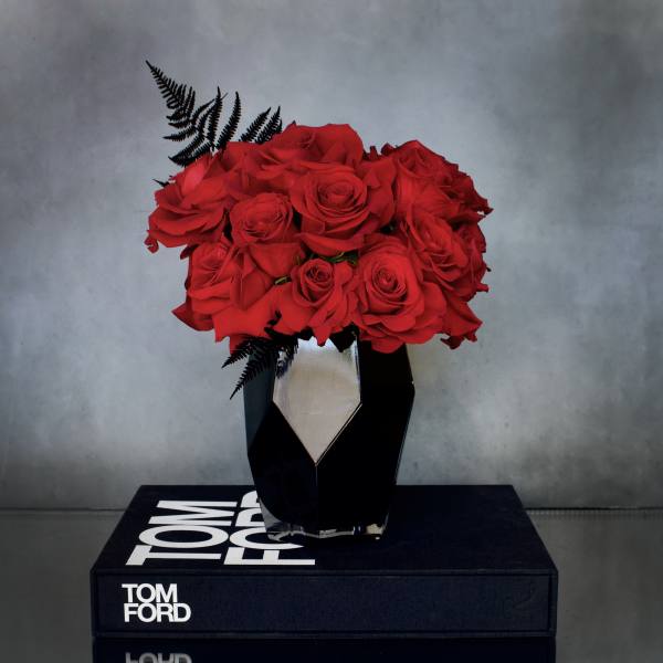 Compact arrangement of red roses with black fern accents in a faceted black vase on a book.