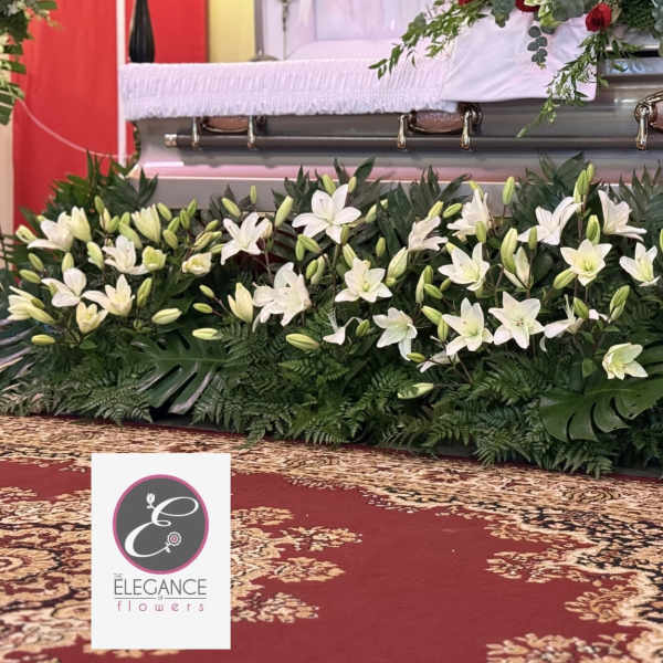 Low arrangement of white lilies with abundant greenery displayed in front of a silver casket.