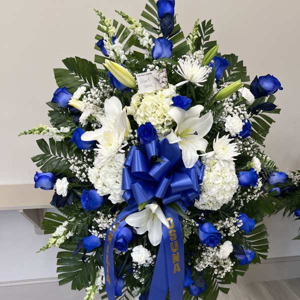Tall standing spray with blue roses and white lilies on an easel with blue ribbon and palm leaves