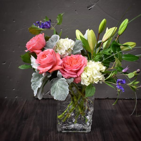 Arrangement of pink roses, white hydrangeas, and lilies in a clear glass cube vase with purple accents