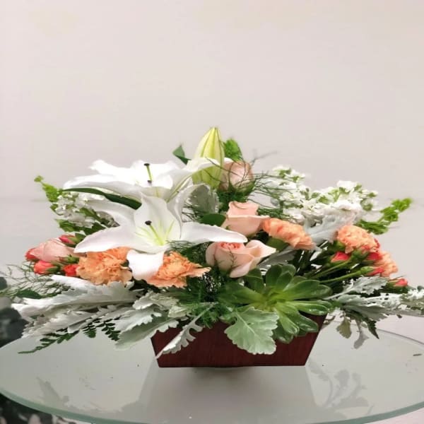 Low floral arrangement with white lilies, pale roses, and peach carnations in a wooden container
