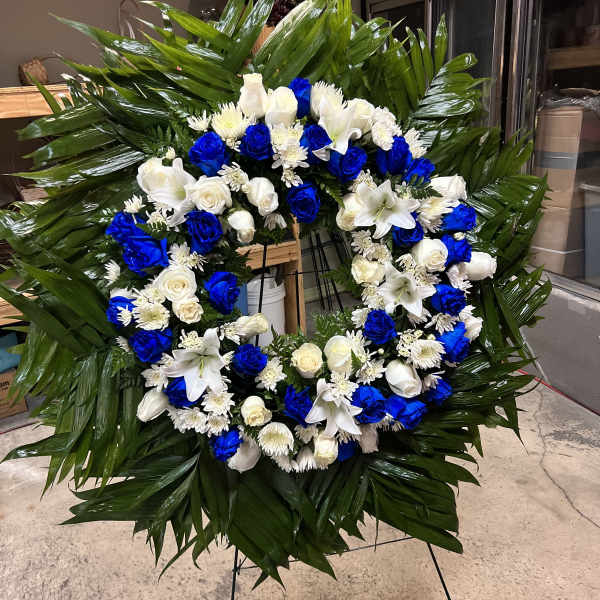 Large standing wreath with blue and white roses, white lilies, and mums on glossy green leaves.