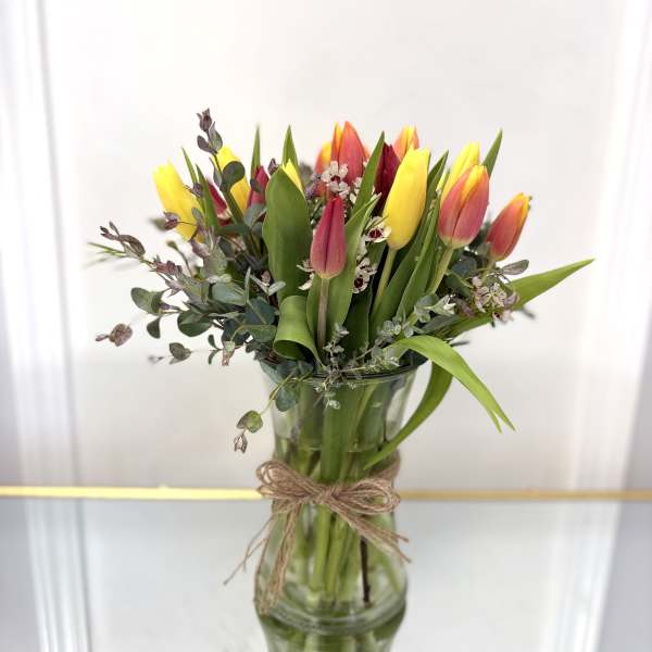 Yellow and red tulips with small white blooms in a tall clear glass vase tied with twine