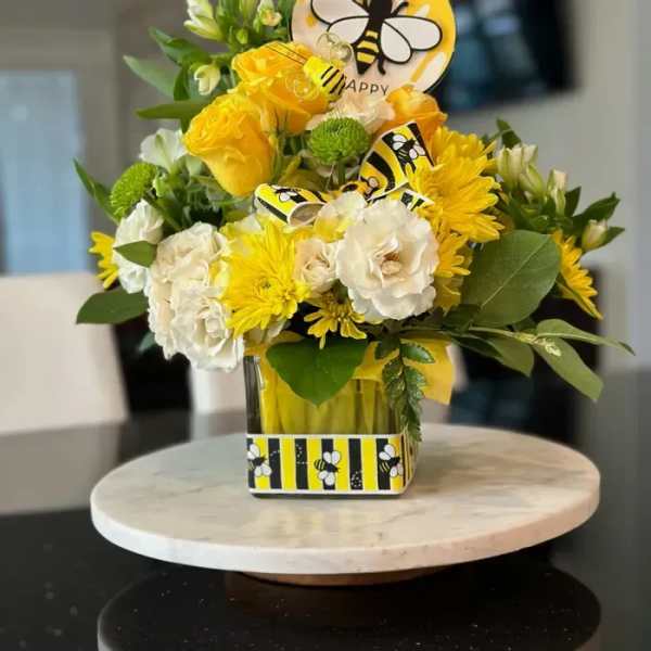 Yellow and white flower arrangement with bee-themed accents in a glass cube vase on a marble stand