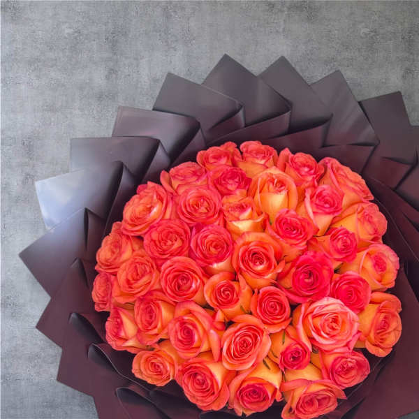 Round bouquet of bright orange and pink roses wrapped in dark folded paper.