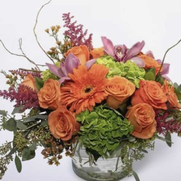 Orange roses and gerbera daisies in a glass vase with purple accents