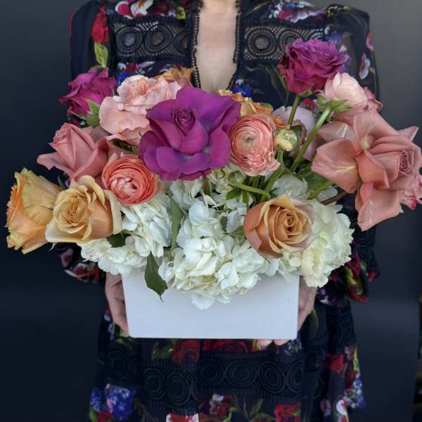 Colorful arrangement of roses and ranunculus with white hydrangeas in a white rectangular box.