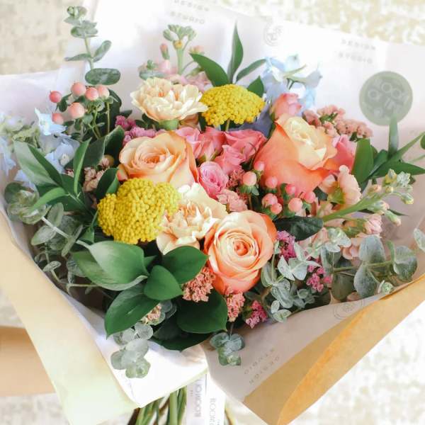 Hand-tied bouquet of peach roses, yellow blooms, and mixed pastel flowers in kraft and white wrap.