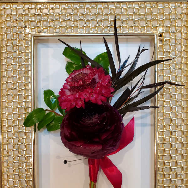 Small floral boutonniere with dark red blooms and a pink ribbon in a gold frame