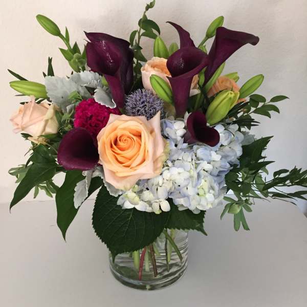Bouquet of peach roses, purple calla lilies, and blue hydrangeas in a glass vase