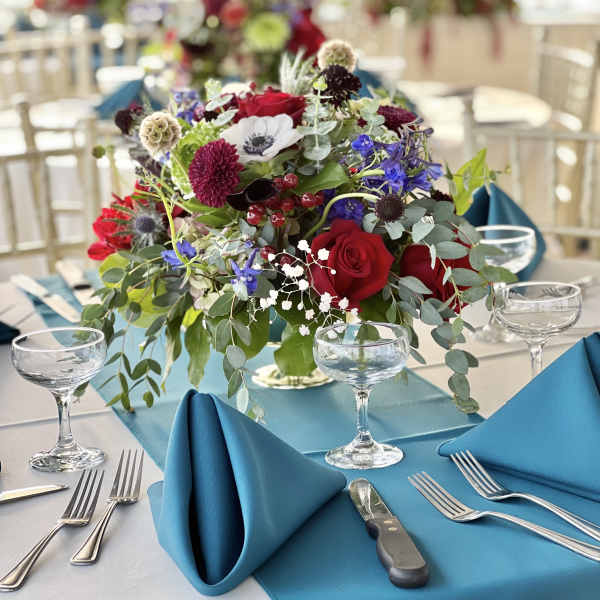 Low centerpiece with red roses, blue flowers, and greenery on a banquet table