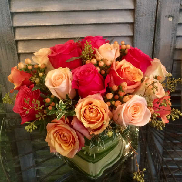 Compact arrangement of pink and peach roses with berries in a square glass vase