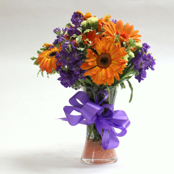 Orange and purple mixed bouquet in a clear glass vase with a purple ribbon