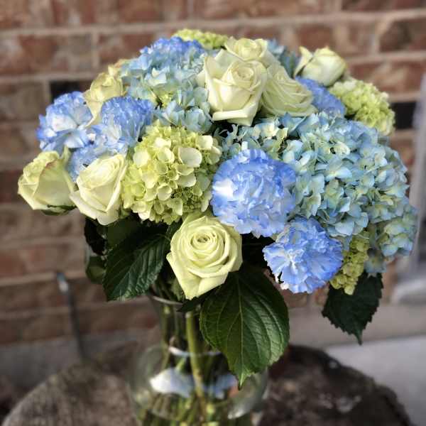 Tall glass vase of pale yellow roses and blue hydrangeas with blue carnations.