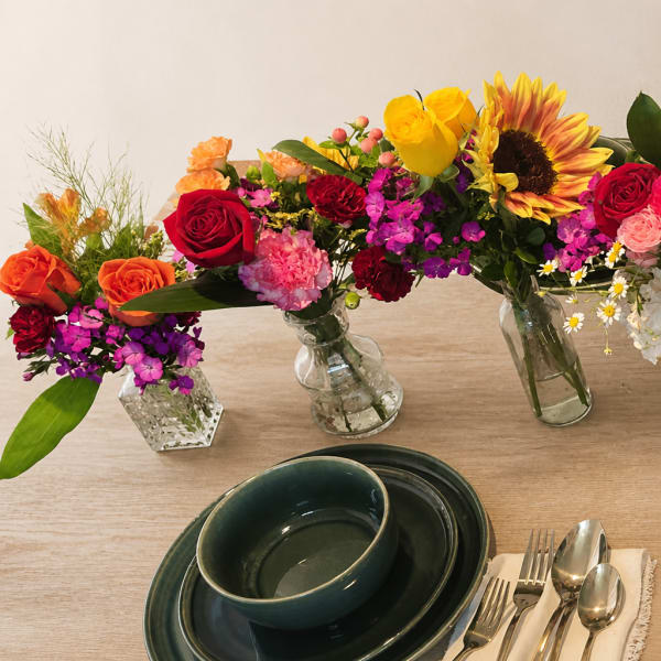 Three colorful flower arrangements in glass vases on a table with dark green dishes