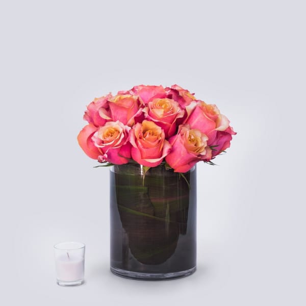 Pink and peach roses arranged in a dark glass vase beside a small candle.