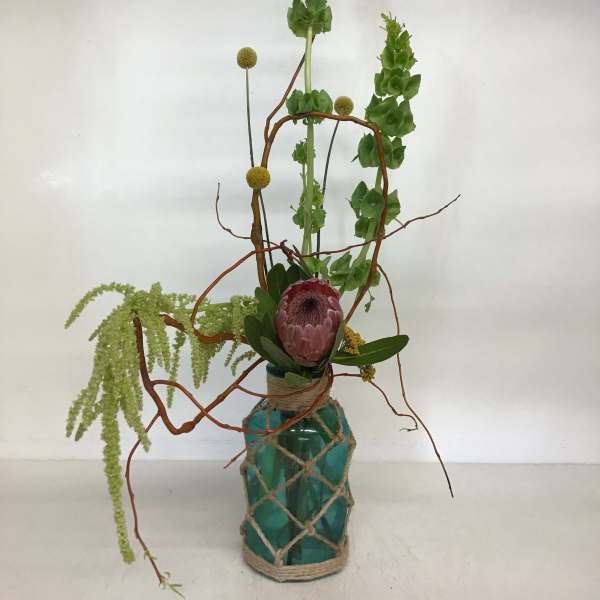 Tall floral arrangement in a teal glass vase with a pink protea and green blooms