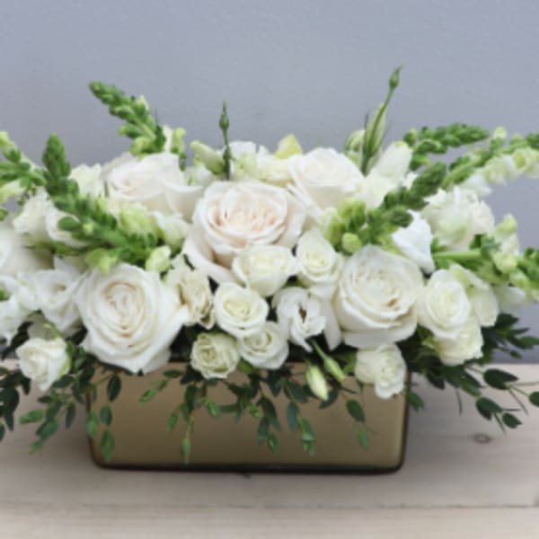 Low rectangular arrangement of white roses and snapdragons in a gold container