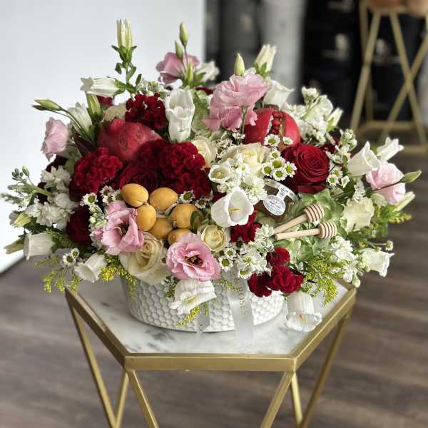 Mixed red, pink, and white floral arrangement in a white bowl with pomegranates and honey dippers