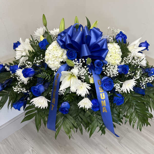 Large blue and white funeral spray with blue roses, white lilies, and a blue bow on a bed of dark foliage.