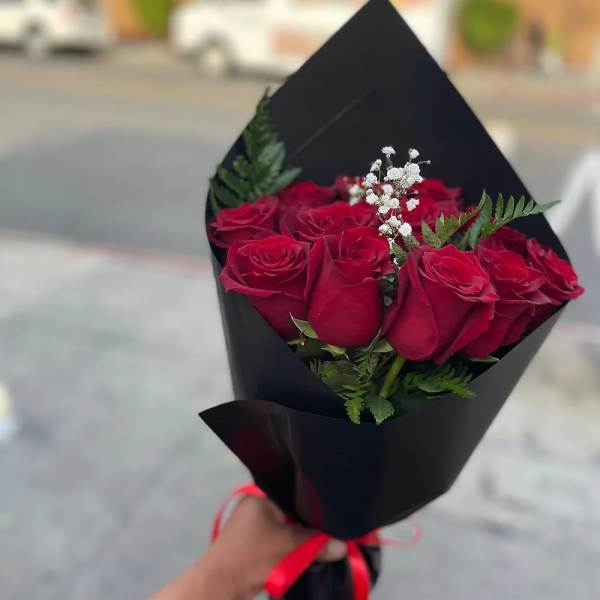 Handheld bouquet of red roses with white filler flowers wrapped in black paper and red ribbon