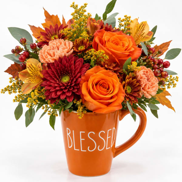 Orange and red floral arrangement in a mug-shaped vase with "BLESSED" text