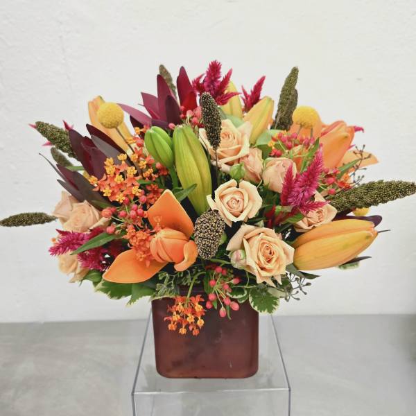 Mixed bouquet of peach roses, orange lilies, and pink accents in a square vase