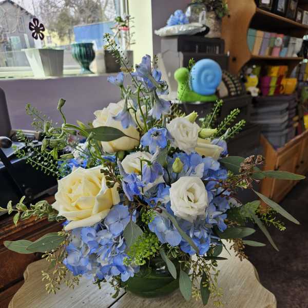 Low arrangement of white roses and blue flowers in a round green glass vase on a small table.