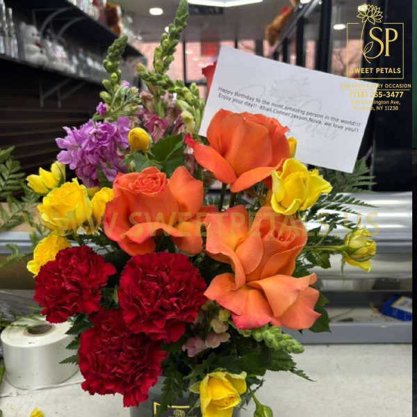 Colorful bouquet with orange roses, yellow roses, red carnations, and purple flowers in a black container.