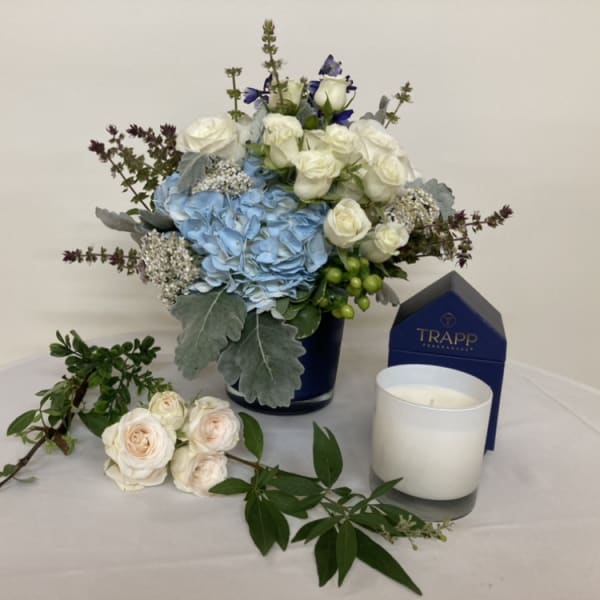 Blue hydrangea and white rose arrangement in a dark vase with a candle and box