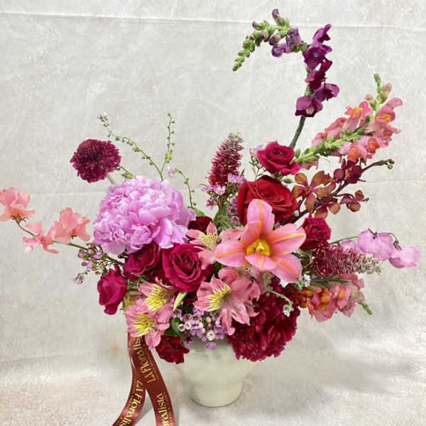Pink and red mixed flower arrangement in a white vase with a ribbon