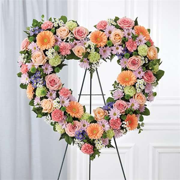 Heart-shaped standing wreath of pink roses, peach gerberas, and pastel daisies on a metal easel.