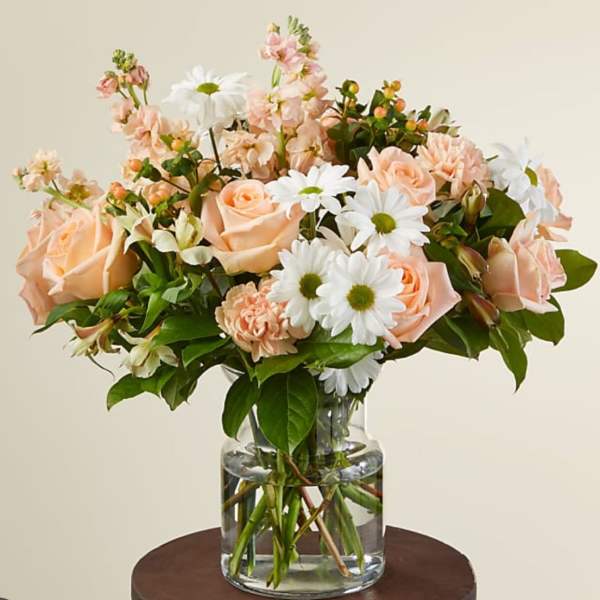 Peach roses, white daisies, and pastel blooms arranged in a clear glass vase.