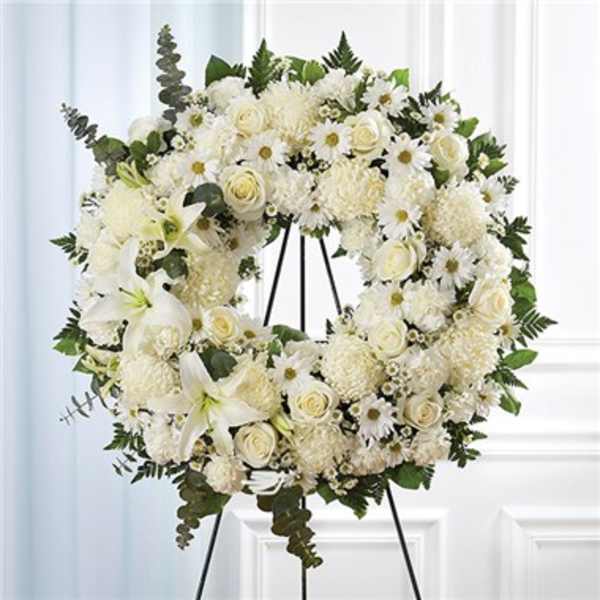 White floral wreath on a black stand with lilies, roses, and daisies