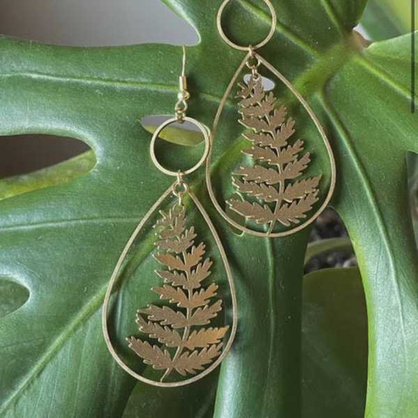 Gold teardrop earrings with fern cutouts on a large green leaf