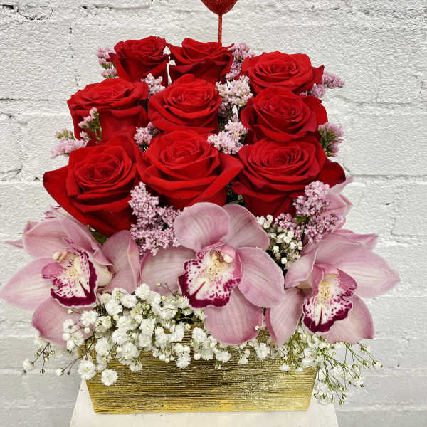Red roses and pink orchids in a gold rectangular container with a heart pick