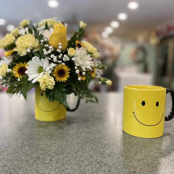 Yellow and white flower arrangement in a smiley-face mug beside a matching mug