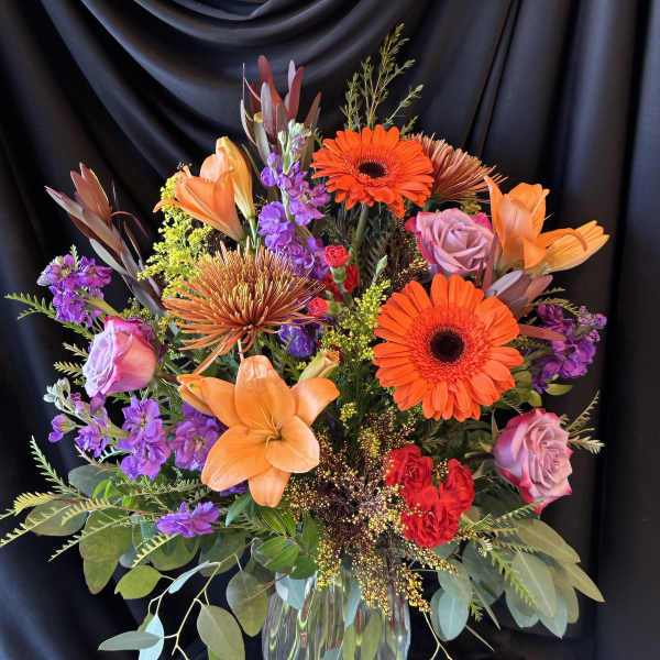 Bright mixed bouquet with orange lilies, gerbera daisies, and purple roses in a glass vase