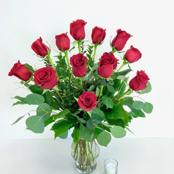Red roses arranged in a clear glass vase with greenery and a small candle beside it
