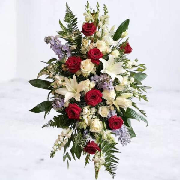 Tall standing spray of red and white roses, white lilies, and soft lavender blooms framed by ferns.