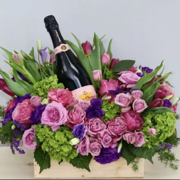 Pink and purple flower arrangement with a champagne bottle in a wooden box
