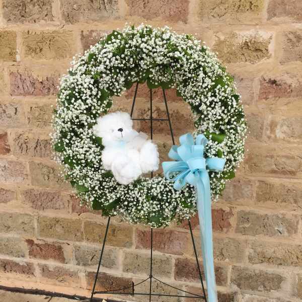 White baby’s breath wreath with white teddy bear and blue bow on a stand against a brick wall