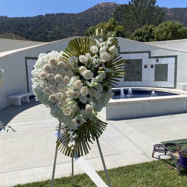 Heart-shaped white rose sympathy spray with gold palm leaves on an easel outdoors.