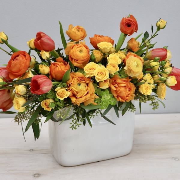 Rectangular white vase filled with yellow spray roses, orange ranunculus, and red-orange tulips.