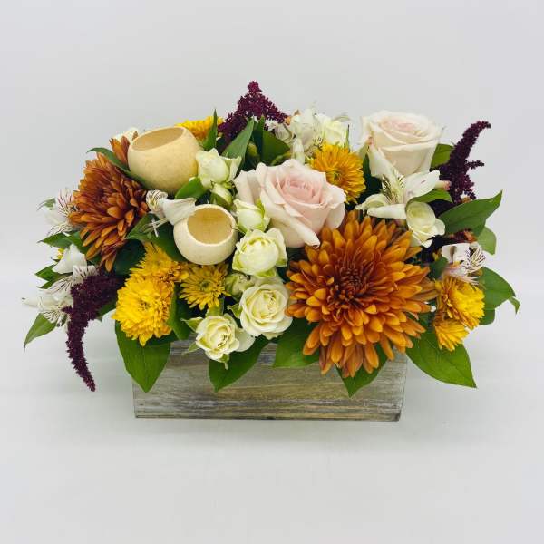 Low rectangular arrangement with orange and yellow mums, pale roses, and white blooms in a wooden box with decorative pods.