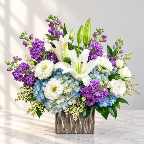 Purple and white floral arrangement with lilies and blue hydrangeas in a metallic vase