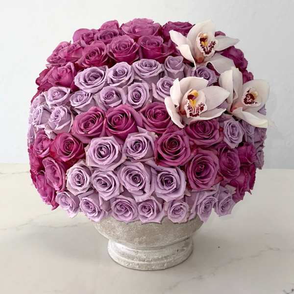 Round bouquet of pink and lavender roses with white orchids in a pedestal vase